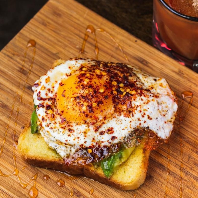 Spicy fried egg on avocado toast with chili flakes and honey drizzle on a wooden board.