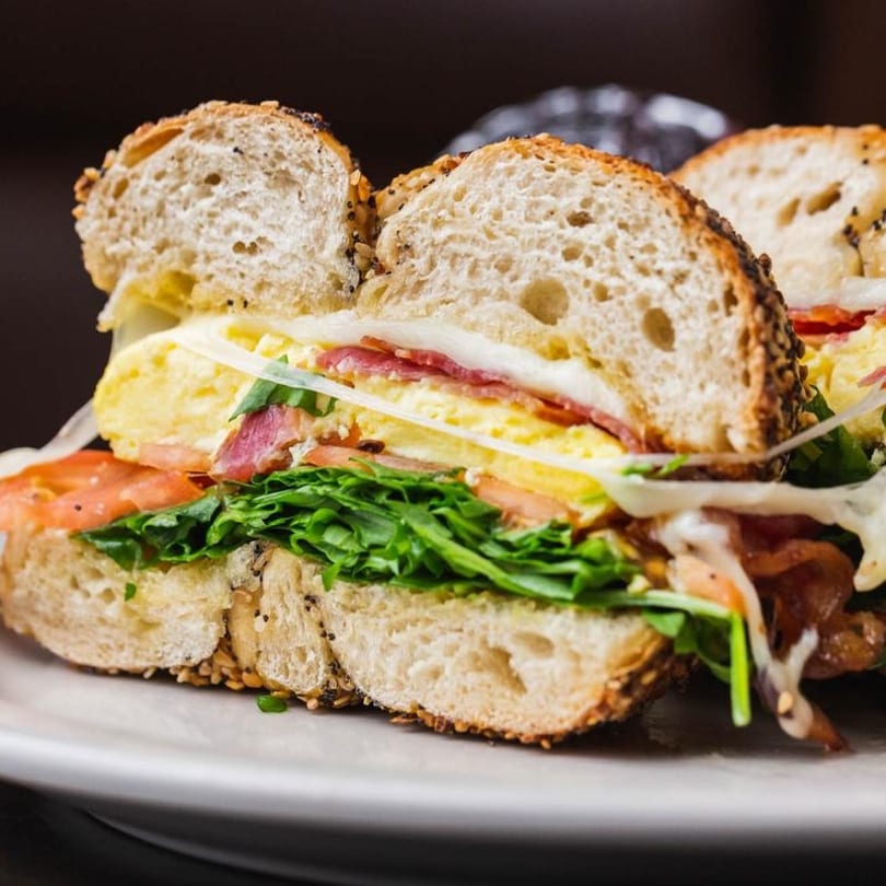 Close-up of a bagel sandwich with egg, bacon, cheese, lettuce, and tomato on a white plate.