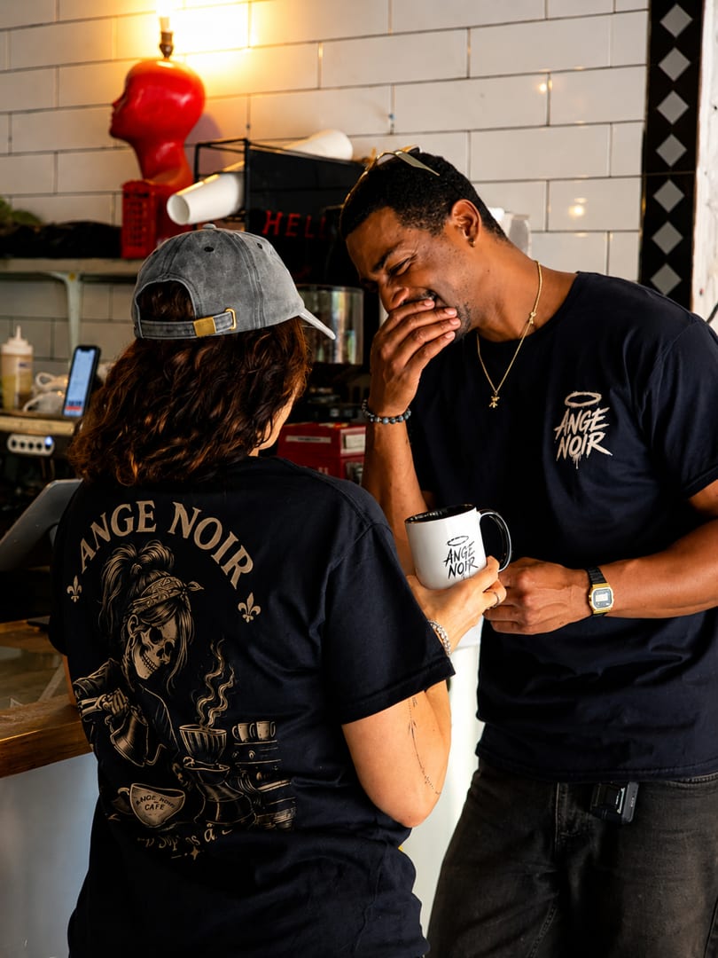 Two people in Ange Noir Cafe shirts laughing and talking at a coffee shop counter.