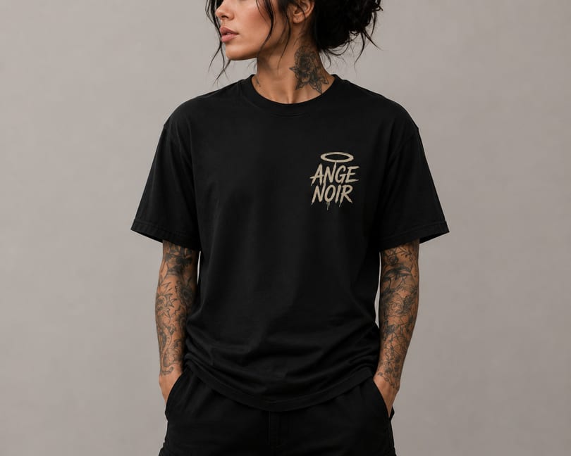 Person with neck and arm tattoos wearing a black t-shirt with "ANGE NOIR" and a halo graphic.