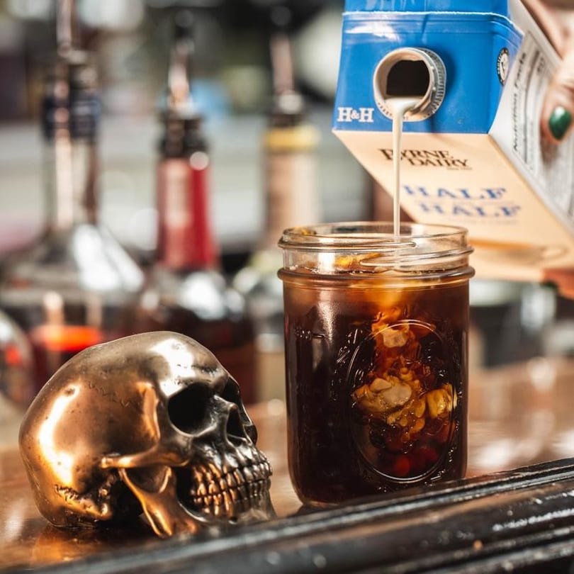 Half and half being poured into a mason jar of iced coffee next to a bronze skull.