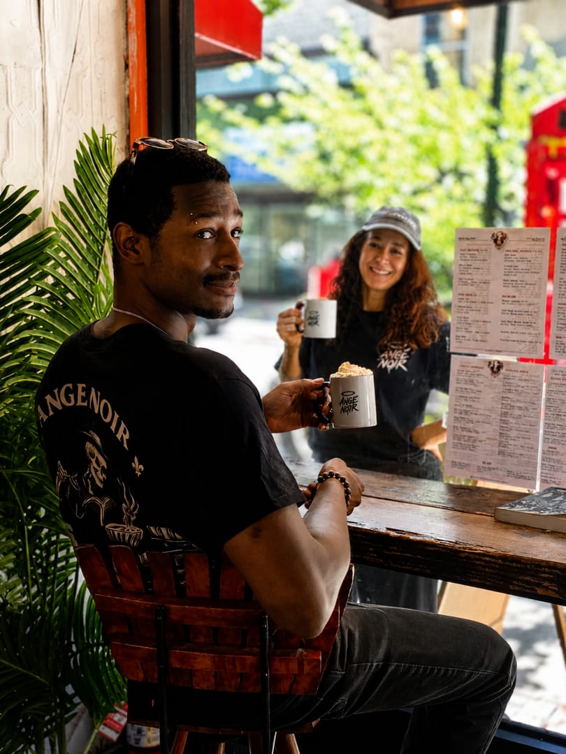 Man in black Ange Noir shirt holding a mug, smiling at the camera, with a woman behind him.