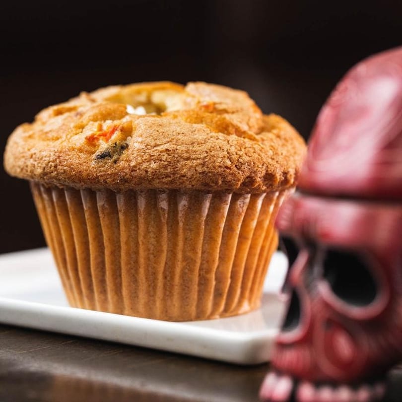 A golden-brown muffin with visible fruit pieces sits on a white plate next to a red skull decoration.